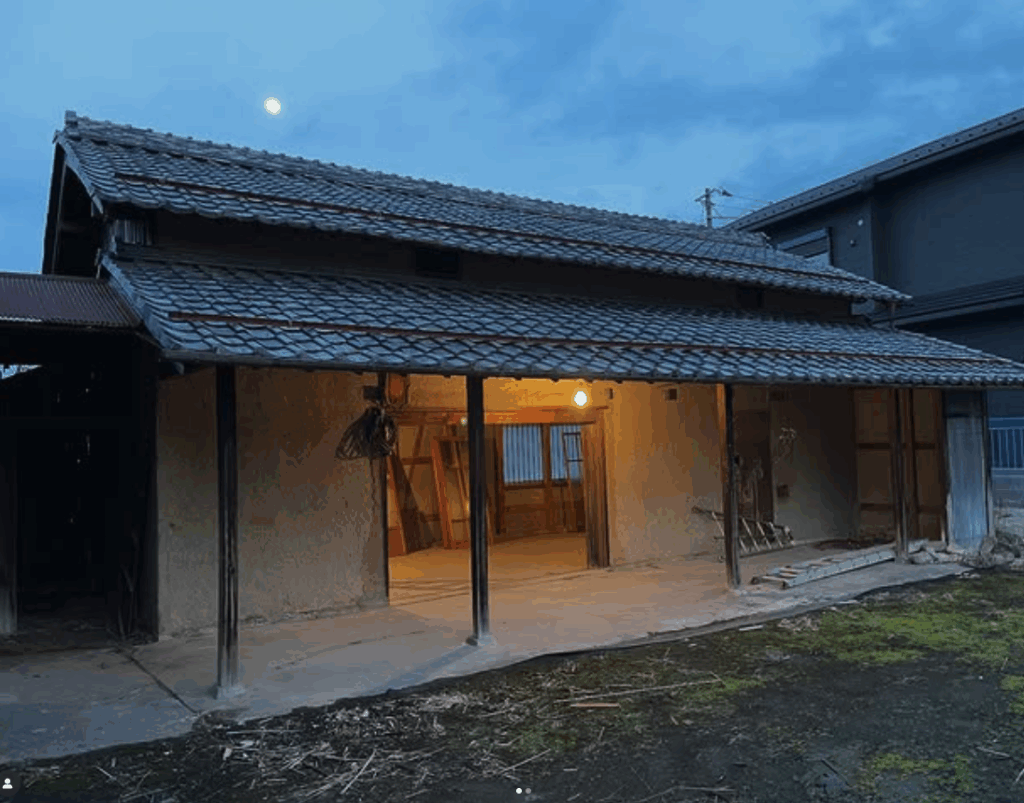 A traditional Japanese house with a thatched roof, partially illuminated from within, set against a twilight sky featuring a visible moon.
