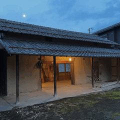 A traditional Japanese house with a thatched roof, partially illuminated from within, set against a twilight sky featuring a visible moon.