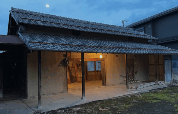 A traditional Japanese house with a thatched roof, partially illuminated from within, set against a twilight sky featuring a visible moon.