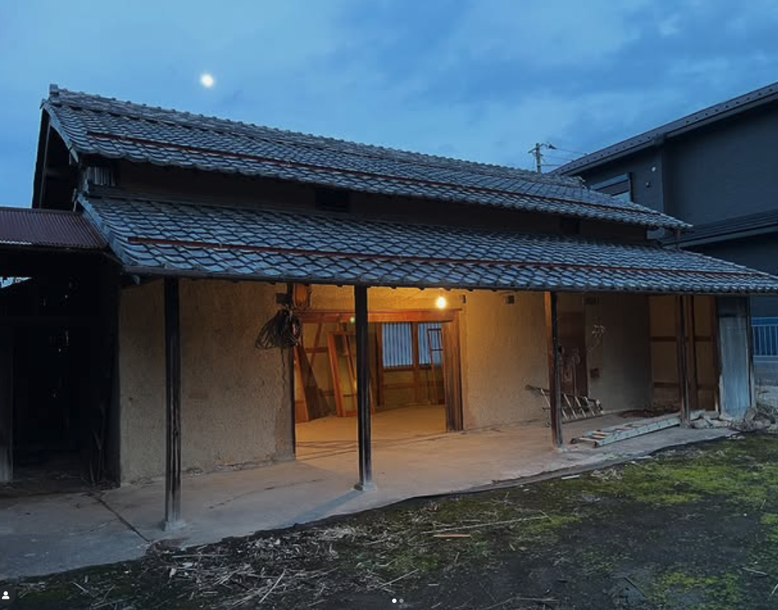 A traditional Japanese house with a thatched roof, partially illuminated from within, set against a twilight sky featuring a visible moon.