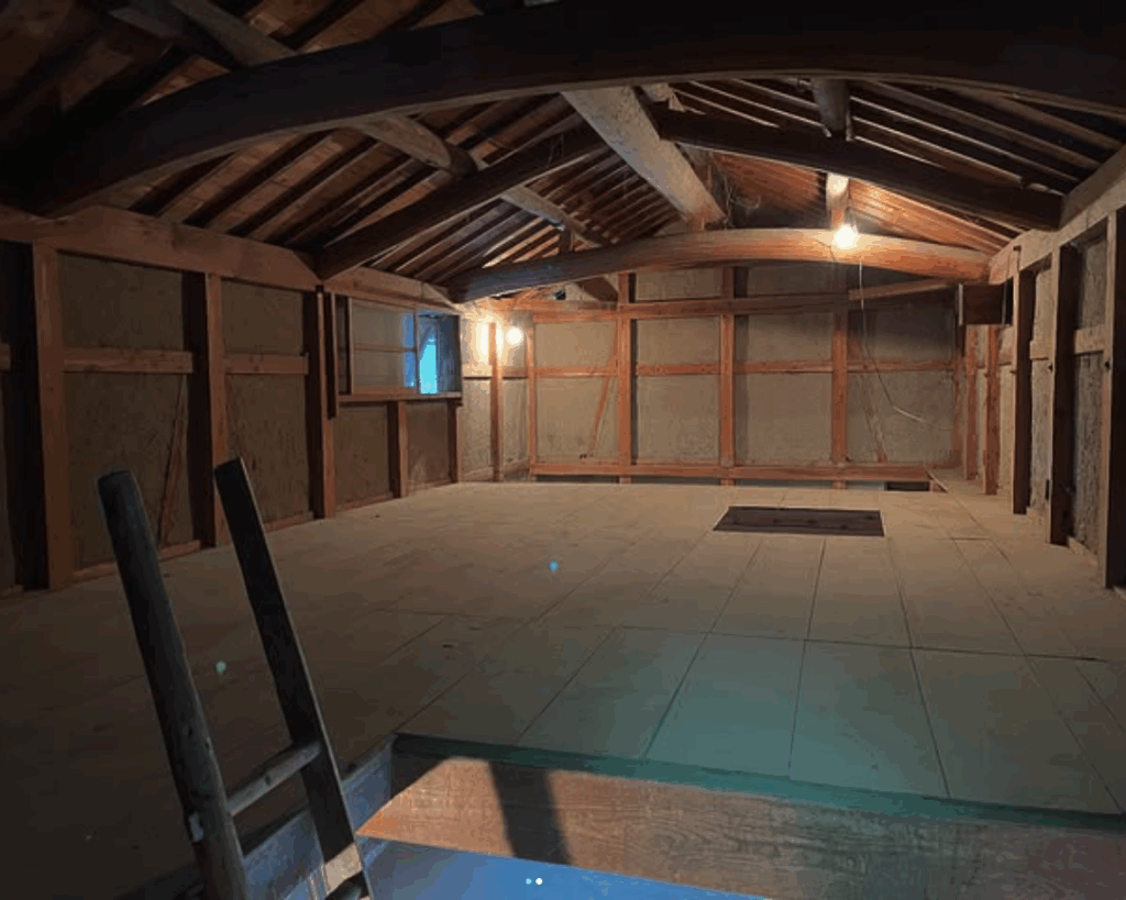 A spacious, rustic attic with wooden beams, a plain floor, and a single light source illuminating the area, showcasing simplicity and potential for use.