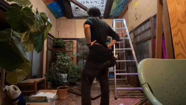 A person adjusts their outfit while standing in a partially renovated room filled with plants, a ladder, and construction materials.
