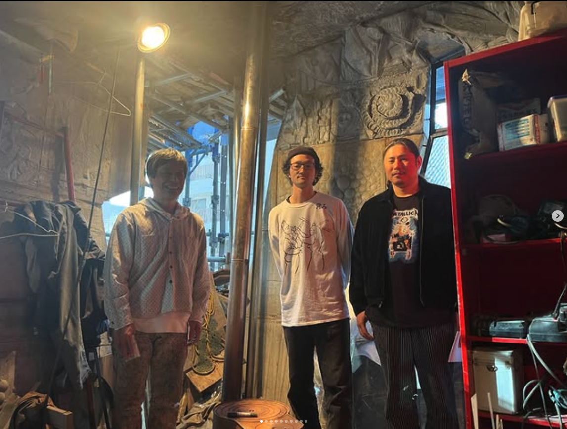 Three individuals standing in a creatively designed workshop, surrounded by tools and art, with warm lighting illuminating the space.