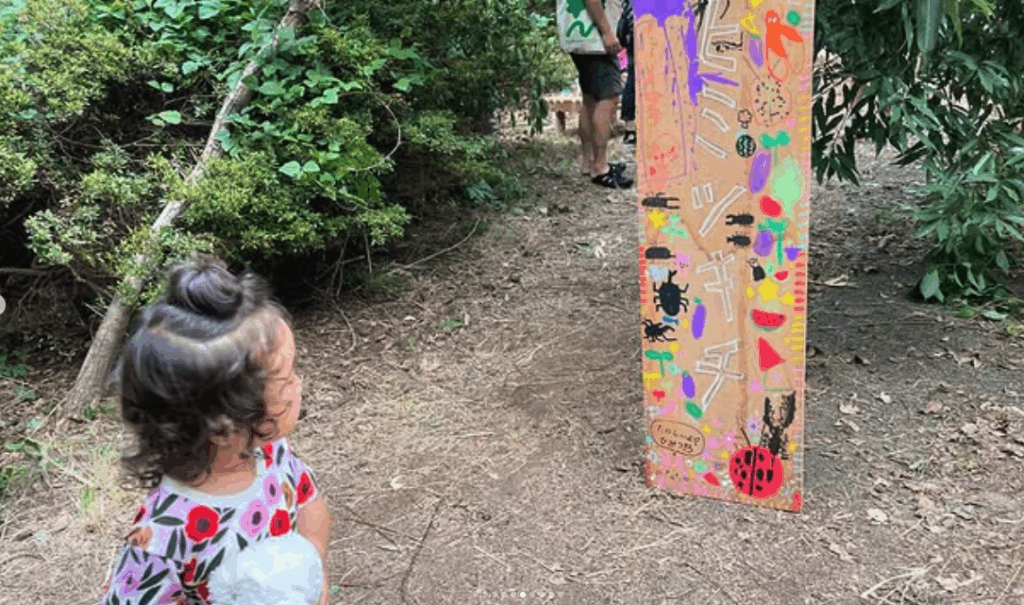 A young child with curly hair observes a colorful sign amidst greenery on a nature trail.